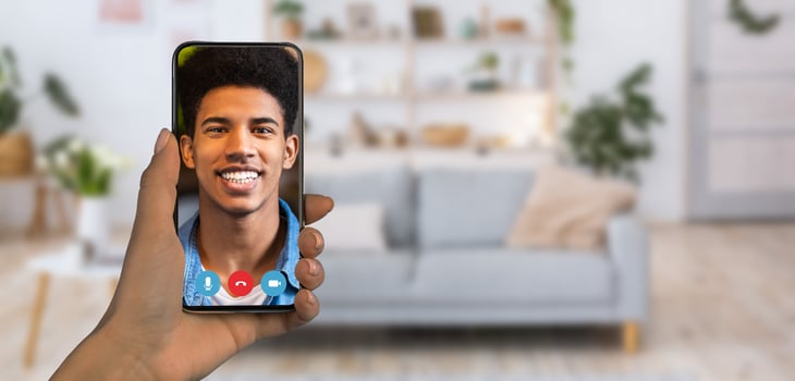A person is holding a smartphone displaying a smiling individual during an online communication session. The cozy home setting features a stylish couch and decorative plants. photo