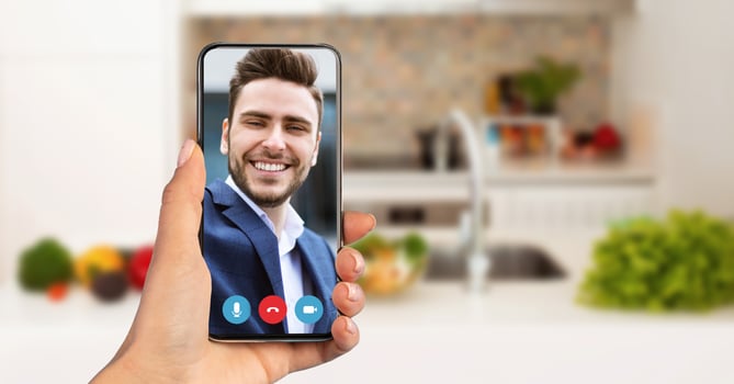 A person holds a smartphone displaying a smiling friend on a call. The setting is a cozy kitchen, filled with fresh produce. The atmosphere suggests a relaxed evening chat. photo