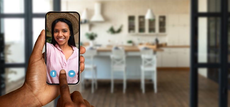 A person holds a smartphone displaying a cheerful individual during a call. The background features a cozy kitchen, indicating a relaxed home atmosphere. photo