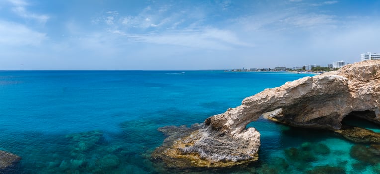 A natural Love arch spans over clear turquoise waters in Ayia Napa, Cyprus, with luxury hotels and resorts visible along the Mediterranean coastline. photo