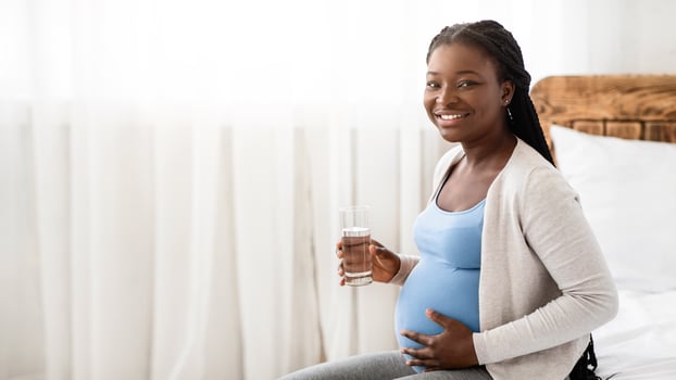 Happy African American Pregnant Woman Drinking Water From Glass While Relaxing On Bed At Home, Smiling Black Expectant Lady Enjoying Healthy Liquid, Thirsty Female Touching Belly And Looking At Camera photo