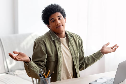 Puzzled black student sitting in front of laptop, not understanding difficult lesson, having difficulty studying online. African American teen guy shrugging shoulders, feeling confused during exam photo