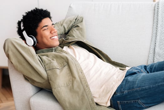 Happy black teen guy in headphones lying on comfy couch and listening to music at home, copy space. African American teenager spending lazy time on weekend, enjoying his favorite song photo