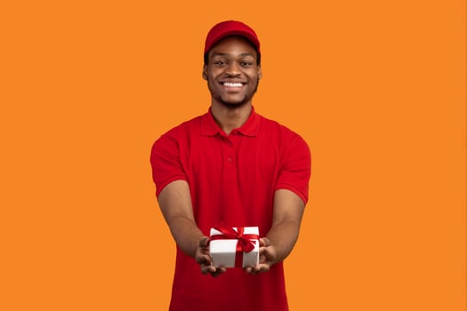 This Present Is For You. Front view portrait of smiling black delivery man holding wrapped gift box giving it to the camera, isolated on orange studio background, greeting with special day photo