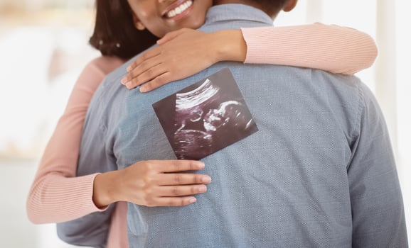Young black pregnant woman with photo of unborn baby hugging her husband indoors, close up. Happy future mother and father with ultrasound picture of their child embracing at home