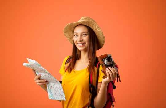 Happy young woman backpacker with camera holding city map over yellow studio background. Joyful lady traveller in summer hat looking at camera and smiling, going for sightseeing abroad, copy space photo