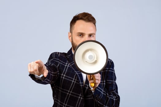 Determined young man in formal wear shouting into megaphone and pointing at you on grey studio background. Public speaker with bullhorn making announcement, proclaiming message photo