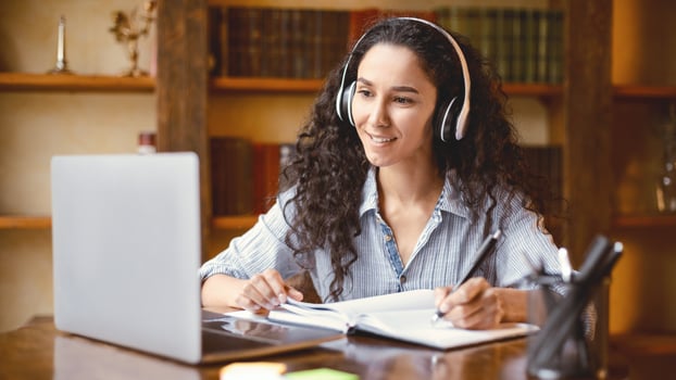 Self Education For Adults. Portrait of beautiful positive young woman wearing wireless headphones watching online course, sitting at table, using personal computer and taking notes in her notebook photo