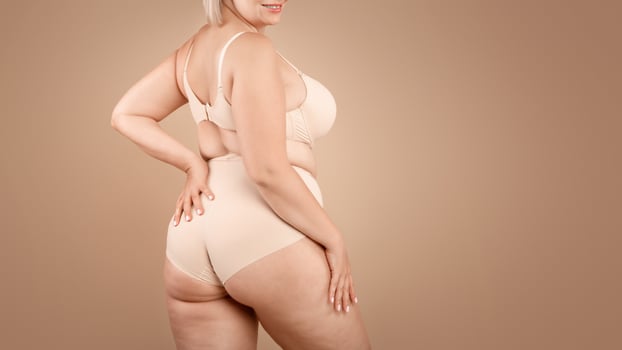 Young plus size woman in beige underwear posing over brown studio background, half-turned at camera, cropped image with free space photo