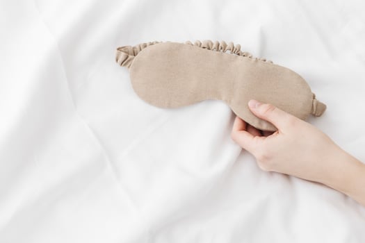 Beige eye mask for sleep on bed background, minimal lifestyle aesthetic, copy space. Top view woman hand holding sleeping mask on white bed linen, morning daylight. Comfort rest, healthy trend photo