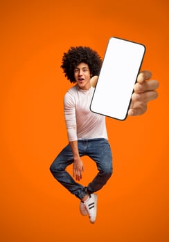 Funny young black teen guy jumping on air, showing cellphone with white empty screen on orange studio background. Young man demonstrating mockup copy space for your mobile app design photo