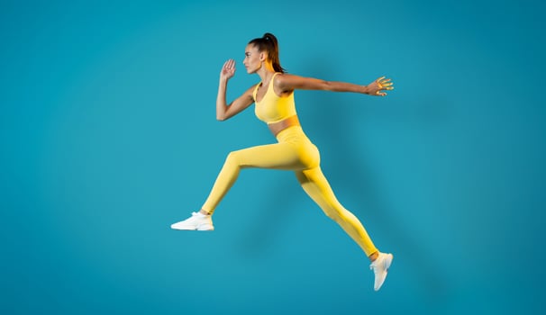 Athletic Woman Jumping Posing In Mid Air Training Over Blue Studio Background. Female Runner Leaping In Air. Workout And Sport Motivation Concept. Side View, Full Length photo