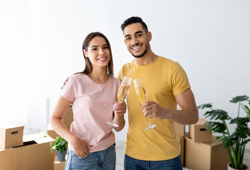 Loving interracial spouses celebrating moving day with champagne, toasting in new apartment among cardboard boxes. Happy millennial married couple moving into their own home photo