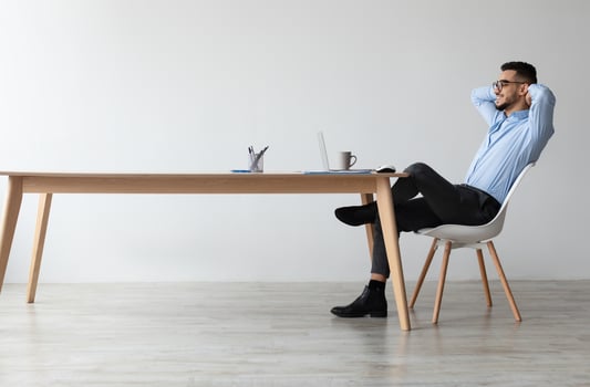 Taking Break. Profile side view of smiling young Arab man relaxing on chair sitting at table and using laptop, happy millennial male leaning back at workplace, looking at pc screen watching photo