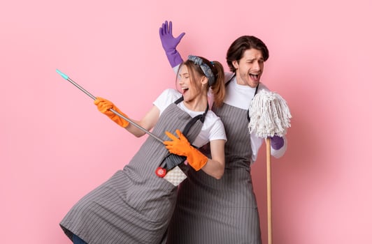 Playful couple of cleaners fooling together while making spring tidying, singing into mop and using broom as guitar over pink background photo