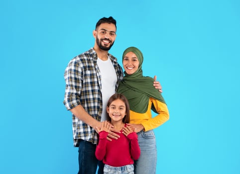 Modern Muslim Family Of Three Hugging Smiling To Camera Standing Together Over Blue Studio Background. Islamic Mother In Hijab And Father Embracing Their Happy Little Daughter photo