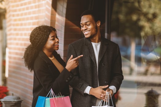 Enjoy shopping on black friday or season sale. Cheerful millennial african american lady with many colored paper bags in hands shows to guy at window and calls to store, free space photo