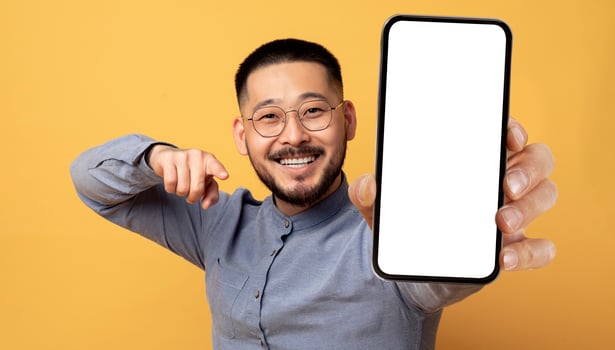 Mobile Ad. Smiling Young Asian Man Pointing At Big Blank Smartphone In His Hand While Standing Over Yellow Studio Background, Happy Millennial Guy Recommending New App Or Website, Collage, Mockup photo
