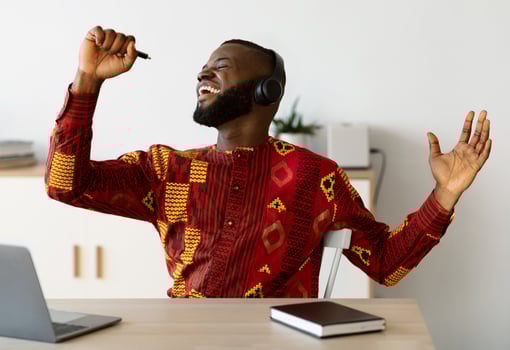 Break In Work. Cheerful African Guy In Traditional Shirt Singing At Workplace, Using Pen As Microphone And Listening Music In Wireless Headphones, Joyful Freelancer Guy Relaxing At Desk In Home Office photo