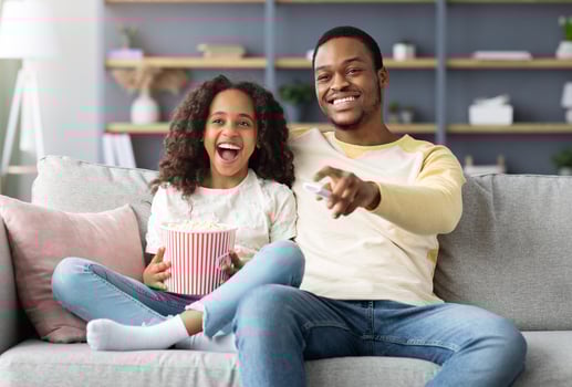 Happy black family father and daughter watching movie together at home. Laughing african american dad and school girl watching funny cartoon and eating popcorn, sitting on couch, copy space photo