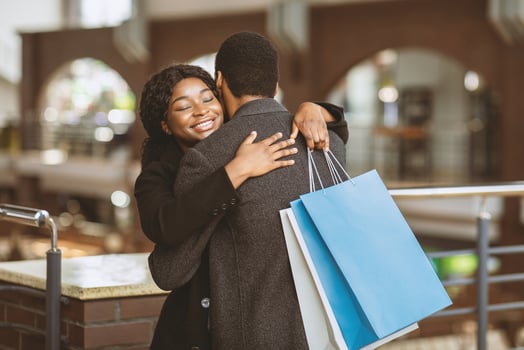 Joyful meeting in store. Happy young african american man and lady in coats hugs in city mall during seasonal sale and Black Friday, female holds colored packages, blurred background, copy space photo