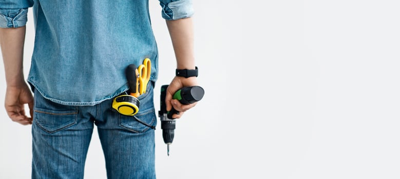Men work, home renovation, construction or carpentry business. Young guy in jeans holds electric drill, with tools in his pocket, isolated on white background, panorama, empty space, cropped, back photo