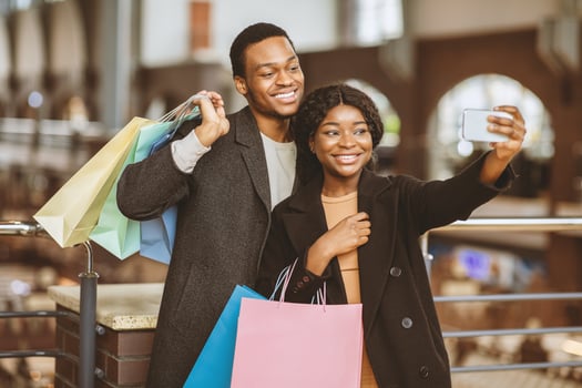 Shopping together during season sale and Black Friday. Happy millennial african american fashionable woman and guy in coats makes selfie at smartphone while walking in city mall, copy space photo