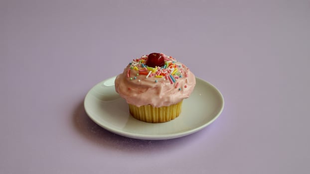 Cupcake with pink frosting and sprinkles topped with a cherry on a white plate on a purple background photo