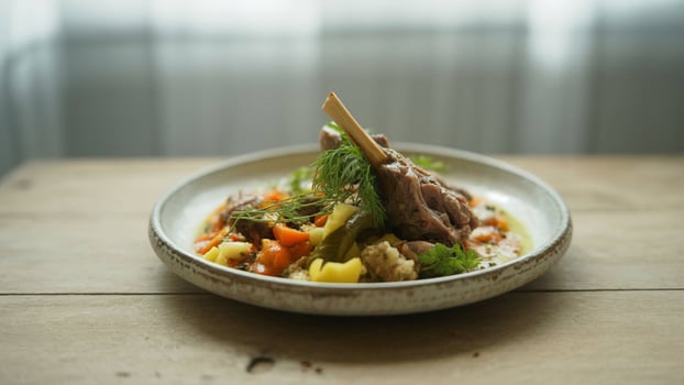 A plate of lamb shank with vegetables and herbs on a wooden table in front of a window light photo