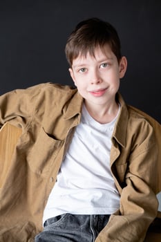 A 12-year-old teenage boy with green eyes in a brown shirt sits on a chair against a black wall, smiling and looking into the frame photo