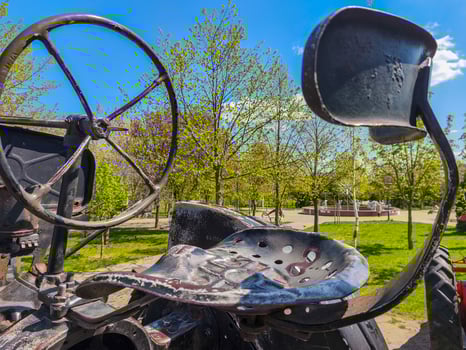 A black tractor with a seat and a steering wheel. The seat is missing and the steering wheel is rusted. The tractor is parked in a park with trees in the background photo