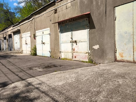 A row of old, abandoned garages sit empty on a street. The buildings are grey and the sky is blue photo