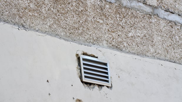 A vent is on the wall of a building. The vent is white and has a black grate photo