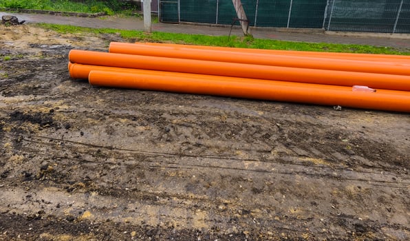 A bunch of orange pipes are laying on the ground. The ground is covered in dirt and there is a fence in the background photo
