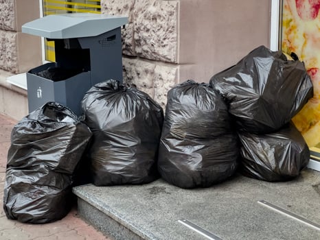 Multiple black trash bags are piled on the sidewalk next to a litter bin in a city setting photo