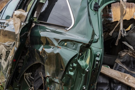 The wrecked green vehicle sits in a rural area, displaying severe dents and torn materials from an accident, surrounded by nature photo