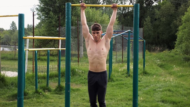 Muscular athlete doing pull ups on horizontal bar at sports ground. Young shirtless sportsman exercising at nature. Strong sporty man training outdoor. Concept of healthy and active lifestyle. Slowmo photo