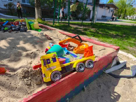 A toy truck is sitting in a sandbox with other toys. The sandbox is filled with sand and has a red wooden fence photo