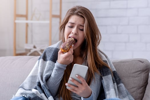 Emotional eating concept. Upset young woman with sweet pastry looking at smartphone, waiting for important message. Anxious millennial lady dealing with stress through comfort food photo