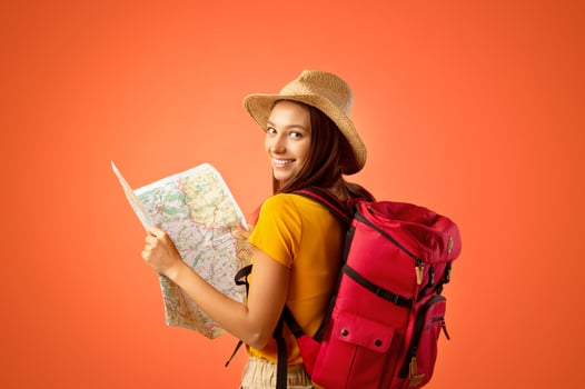 Smiling young woman tourist with backpack holding city map, yellow studio background. Cheerful lady holding map, looking over shoulder, impressed by great travelling opportunities, copy space photo