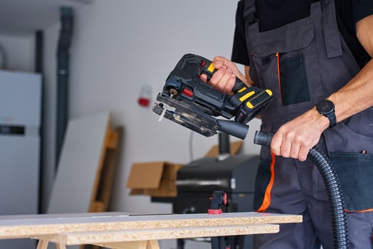 Man in workwear attaching vacuum hose to electric jigsaw for woodworking on workbench in home workshop photo