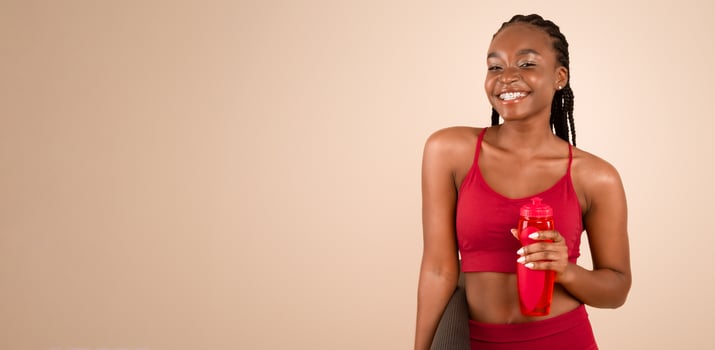 Sporty Lifestyle. Portrait Of Joyful Black Young Woman In Sportswear Holding Fitness Mat And Bottle With Water, Ready For Yoga Training Or Workout, Standing On Beige Studio Background, Copy Space photo