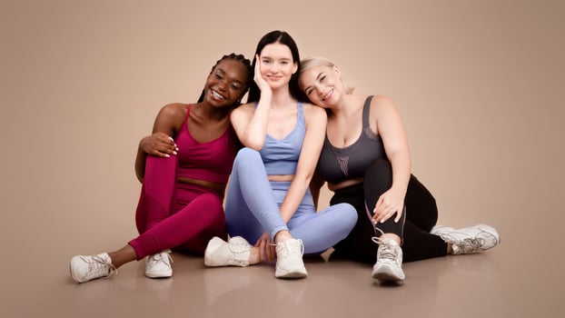 Multi ethnic female friends in sportswear posing in studio, diverse happy women of different size and appearance sitting on floor over beige background and smiling at camera, copy space photo