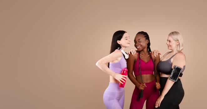 Diverse body positive females in sportswear laughing and posing together, young multi-ethnic ladies standing over beige background in studio, enjoying fitness and healthy lifestyle, copy space photo