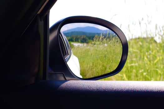 A side mirror reflects a green field and forested hills under a bright sky, captured from inside a car to convey a peaceful travel moment in nature. photo