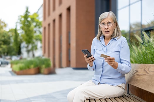 An older woman, seated outdoors, reacts with surprise while looking at her credit card and phone. photo
