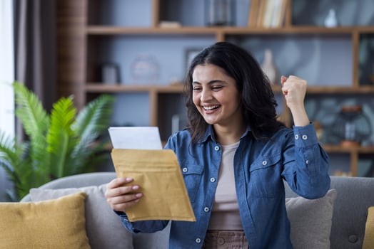 A joyful young woman celebrates good news, reading a letter with a smile and a raised fist in a cozy living room. photo