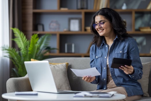 A smiling young woman works from home, holding documents and a tablet while using her laptop on the couch. photo