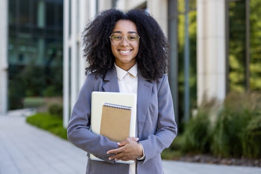 Confident african american business woman in professional attire stands smiling outside modern office building, holding notebook and tablet, embodying success and confidence in workplace. photo