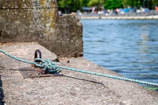 A blue and white rope is tightly knotted to a metal mooring ring on a concrete embankment by a river. photo